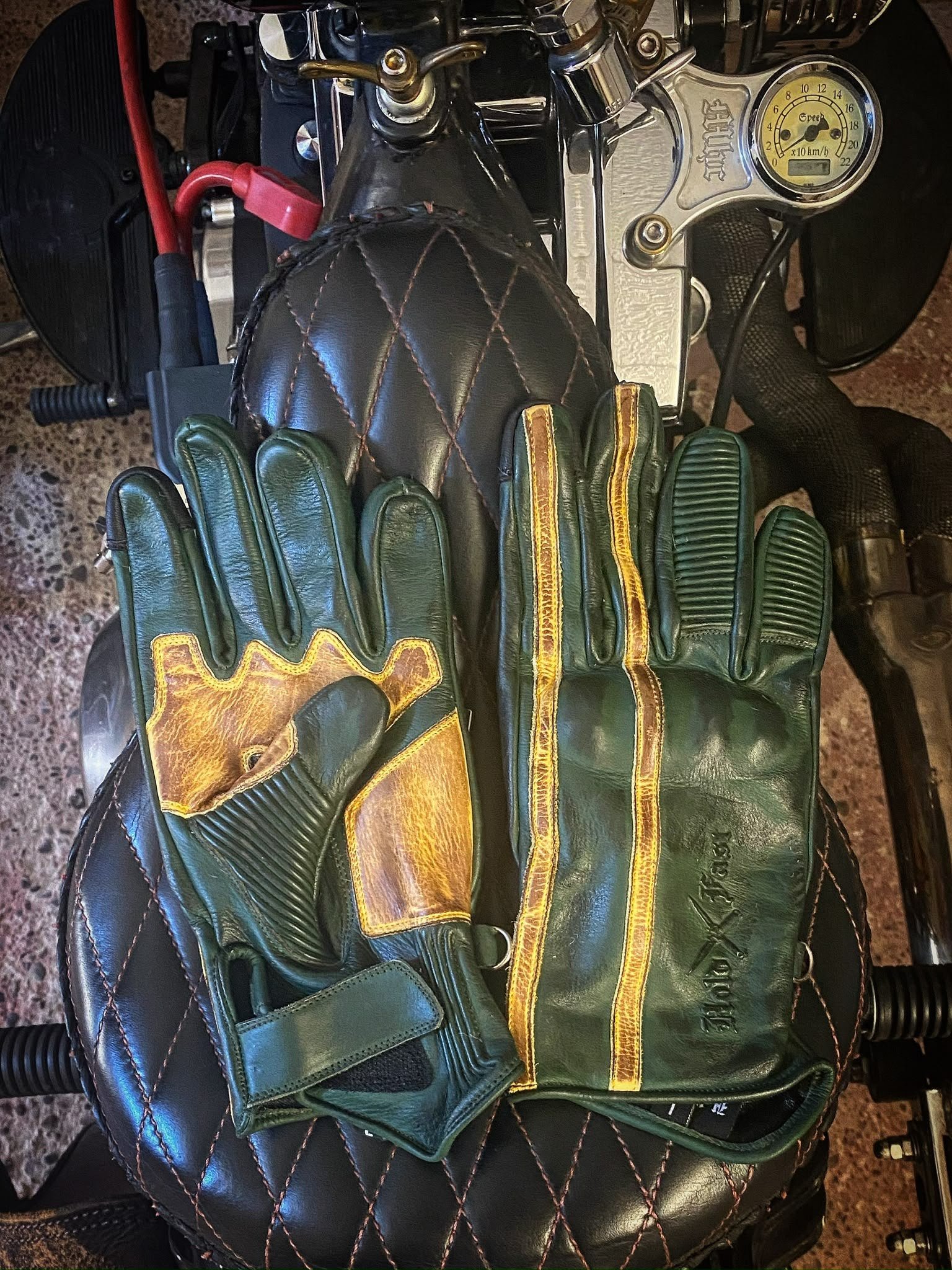 1971 Le Mans Motorcycle Gloves — Green Patina Vintage Racing Hold Fast 1971 Le Mans green patinated leather gloves, showing the palm and back on a quilted motorcycle seat.