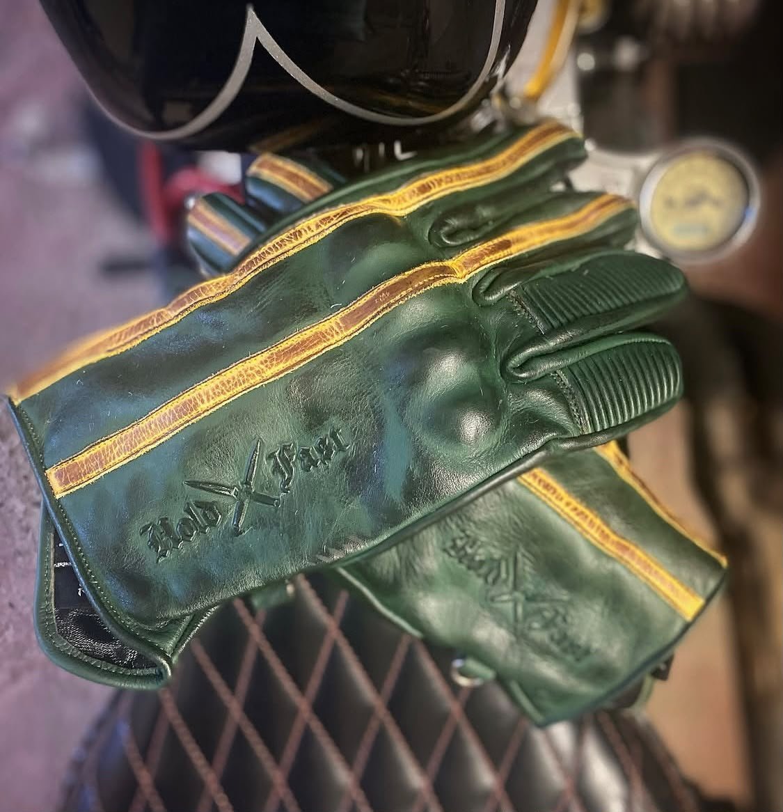 1971 Le Mans Motorcycle Gloves — Green Patina Vintage Racing Close-up of green Hold Fast Le Mans leather gloves with ochre stripes and an embossed logo on a motorcycle seat.